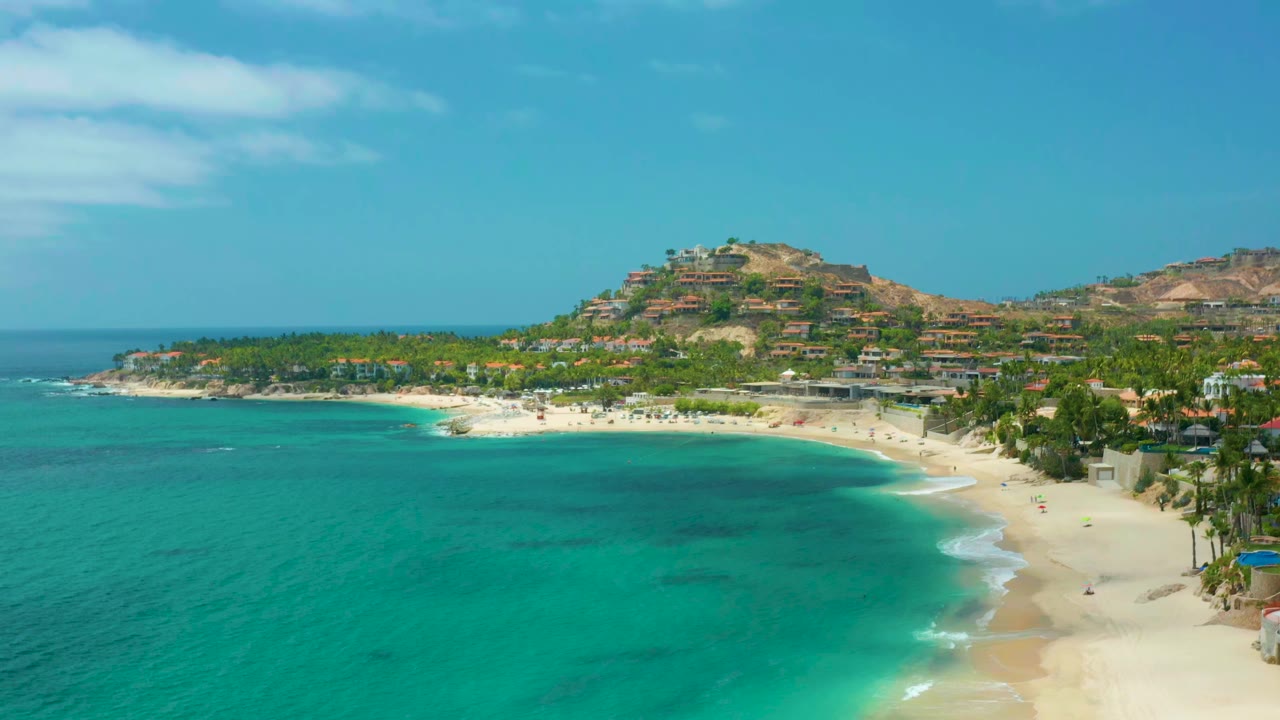 Palmilla Beach Cabo aerial frame with the curved beach more centered, darker patches visible under the turquoise water, hillside buildings in the background, and no visible people.