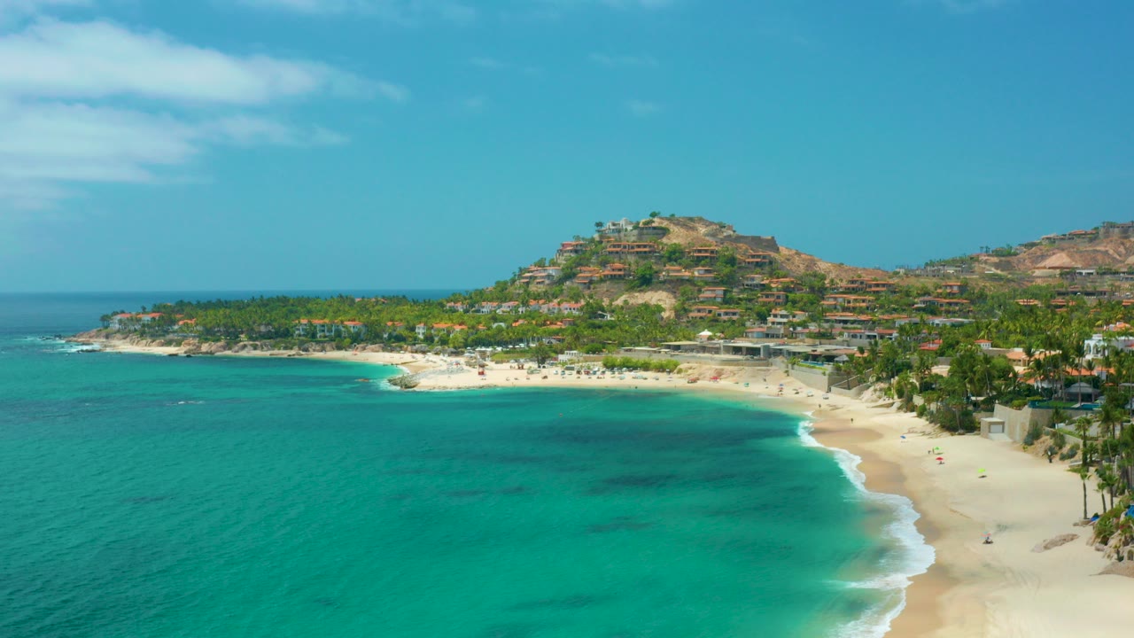 Opening aerial frame over Palmilla Beach Cabo, Mexico, with turquoise sea in front, pale sand curving right, palm trees along the shore, hillside buildings behind, and no visible people.