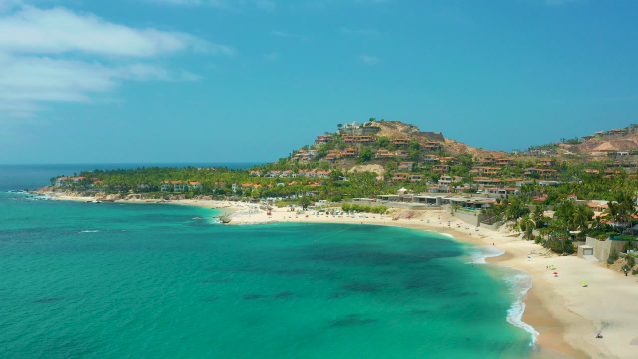 Aerial view at Palmilla Beach Cabo, Mexico with turquoise water in the foreground, a white sand beach at right, palms along the shore, hillside buildings beyond, and tiny people visible on the sand.