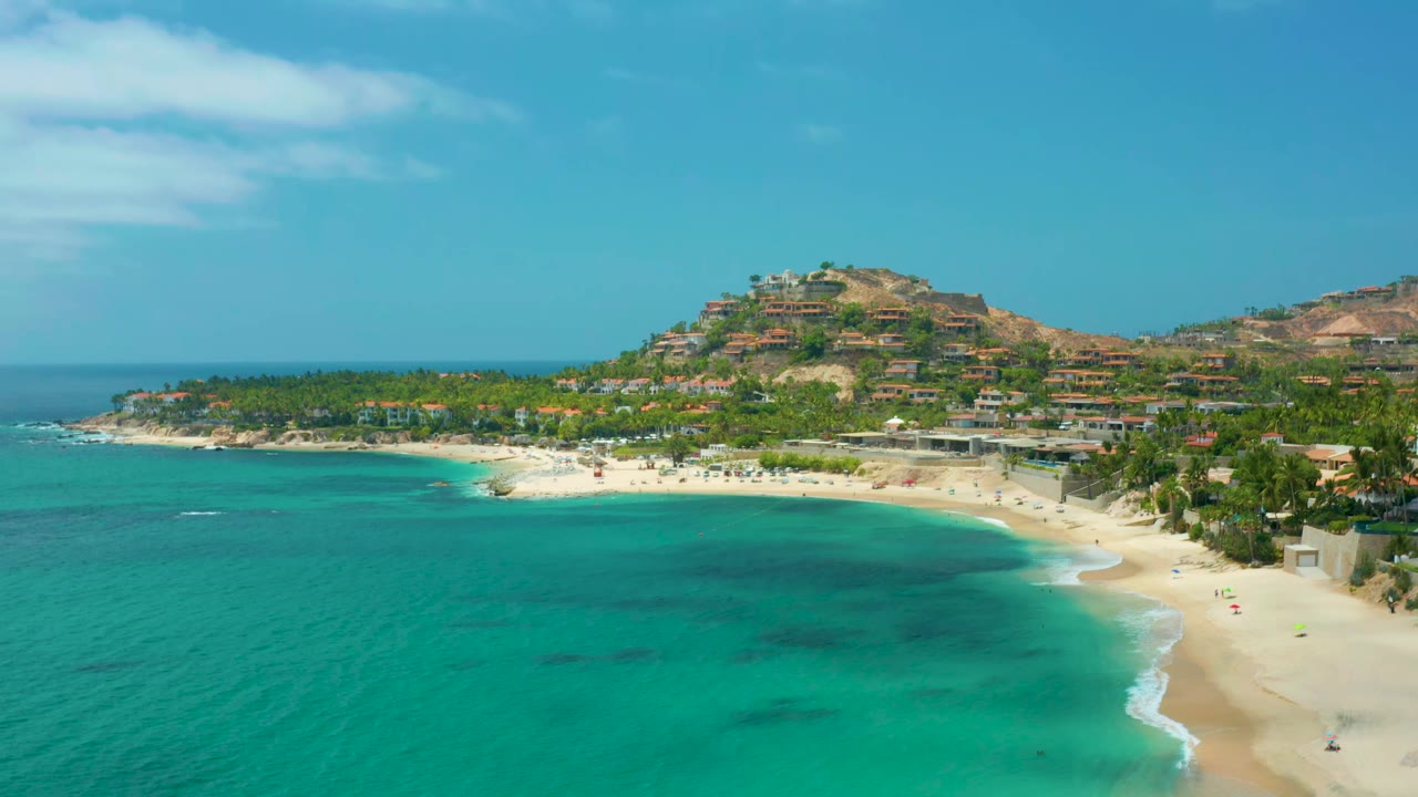 Wide aerial coastline with turquoise sea, pale beach, palm trees, and hillside homes.