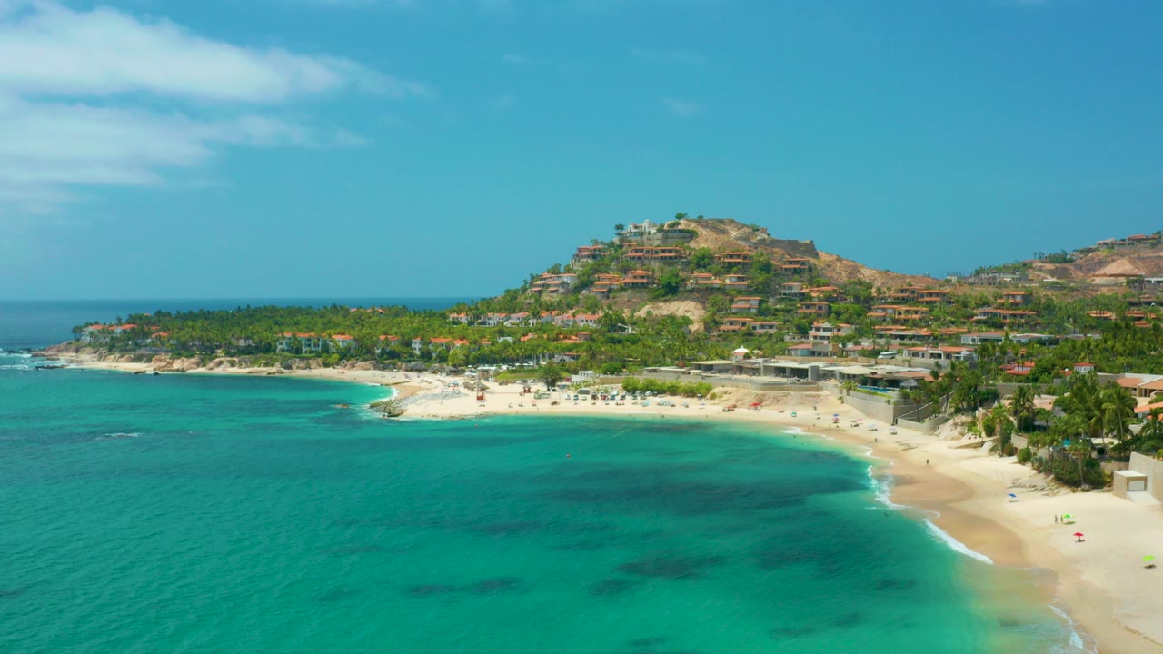 Opening aerial frame over Palmilla Beach Cabo, Mexico, with turquoise water in front, pale sand along the bay, palm trees and hillside buildings beyond, and no visible people.