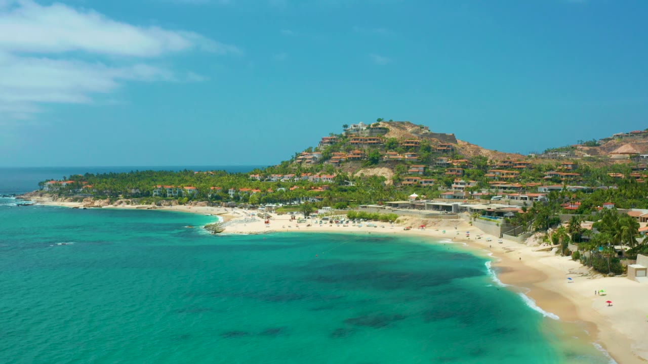 Palmilla Beach Cabo, Mexico seen from above, with teal water darkening in patches, a bright beach strip, palms and low buildings behind it, and no visible people.