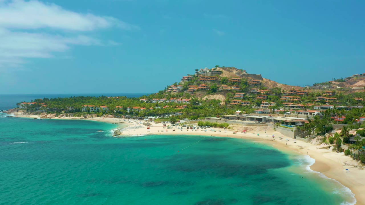 Wide aerial frame of Palmilla Beach Cabo, Mexico showing clear turquoise water, a curving sand beach, a palm-lined shore, hillside homes, and no visible people.