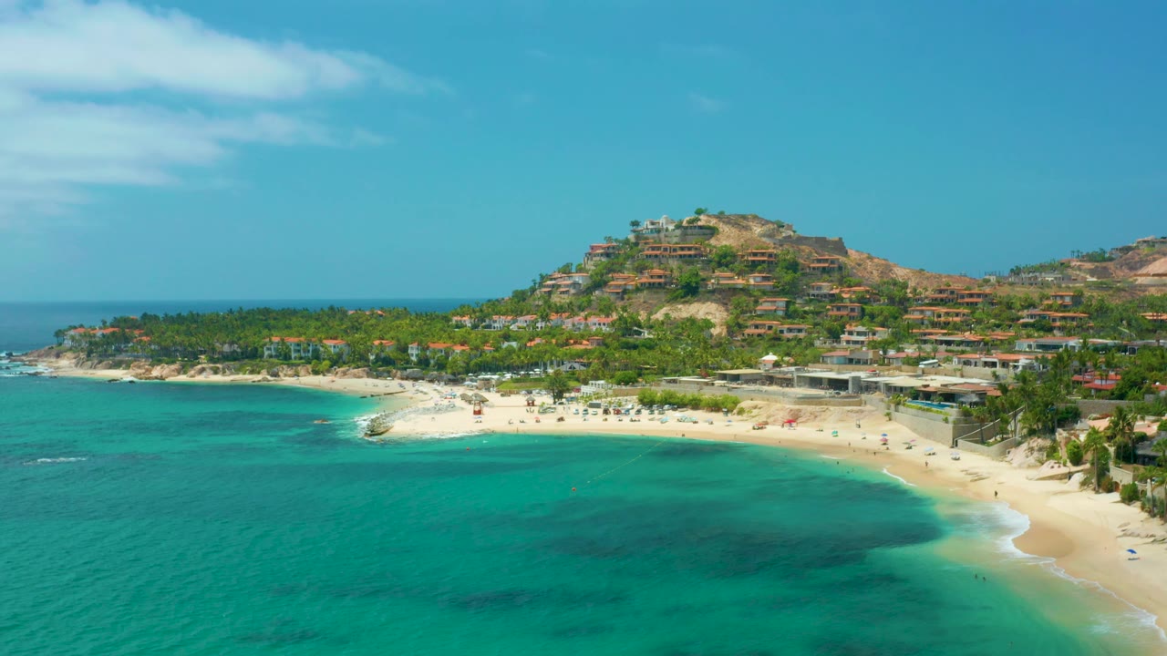 Coastal aerial over Palmilla Beach Cabo, Mexico with the turquoise bay filling the foreground, sand and palms across the middle, hillside development beyond, and no visible people.
