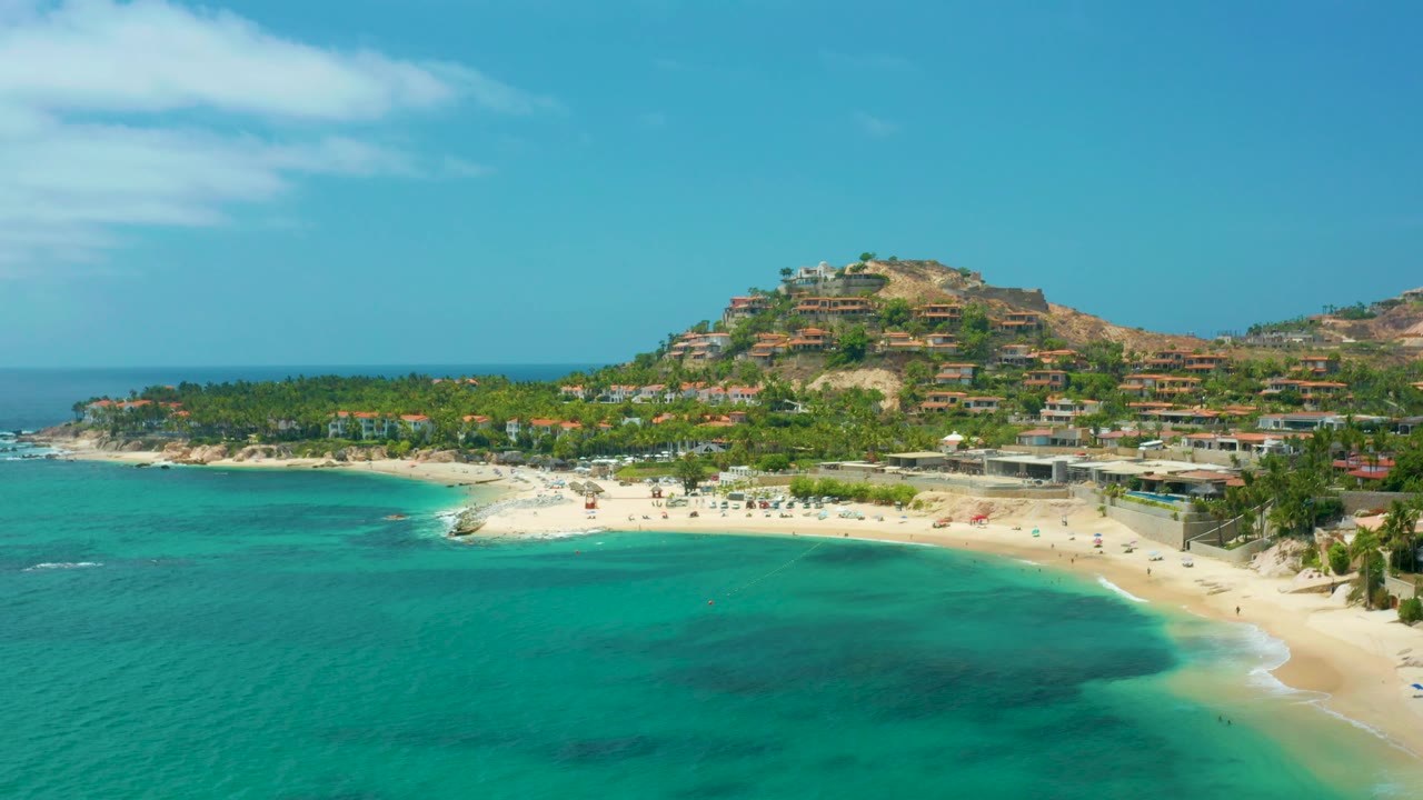 Aerial coastline view with beach, shallow water, palms, and hillside villas.