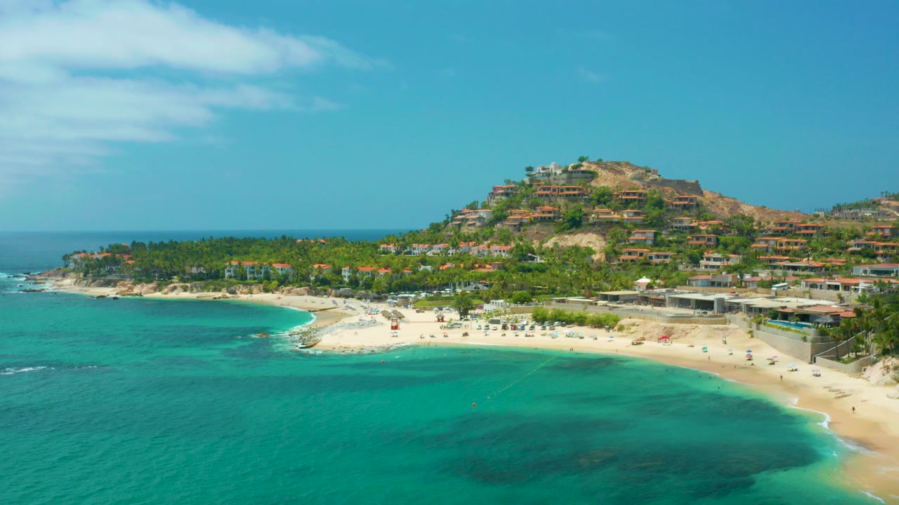 Opening frame of Palmilla Beach Cabo, Mexico with turquoise water in the foreground, a curved sandy beach, palm trees, hillside buildings, and no visible people.