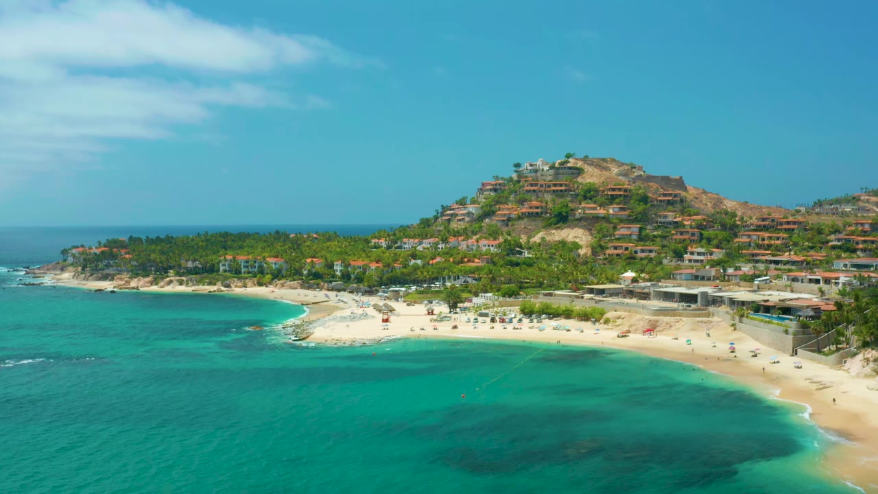 Palmilla Beach Cabo, Mexico seen from above with darker reef-like patches under turquoise water, a pale sand shoreline, palms, hillside buildings, and no visible people.