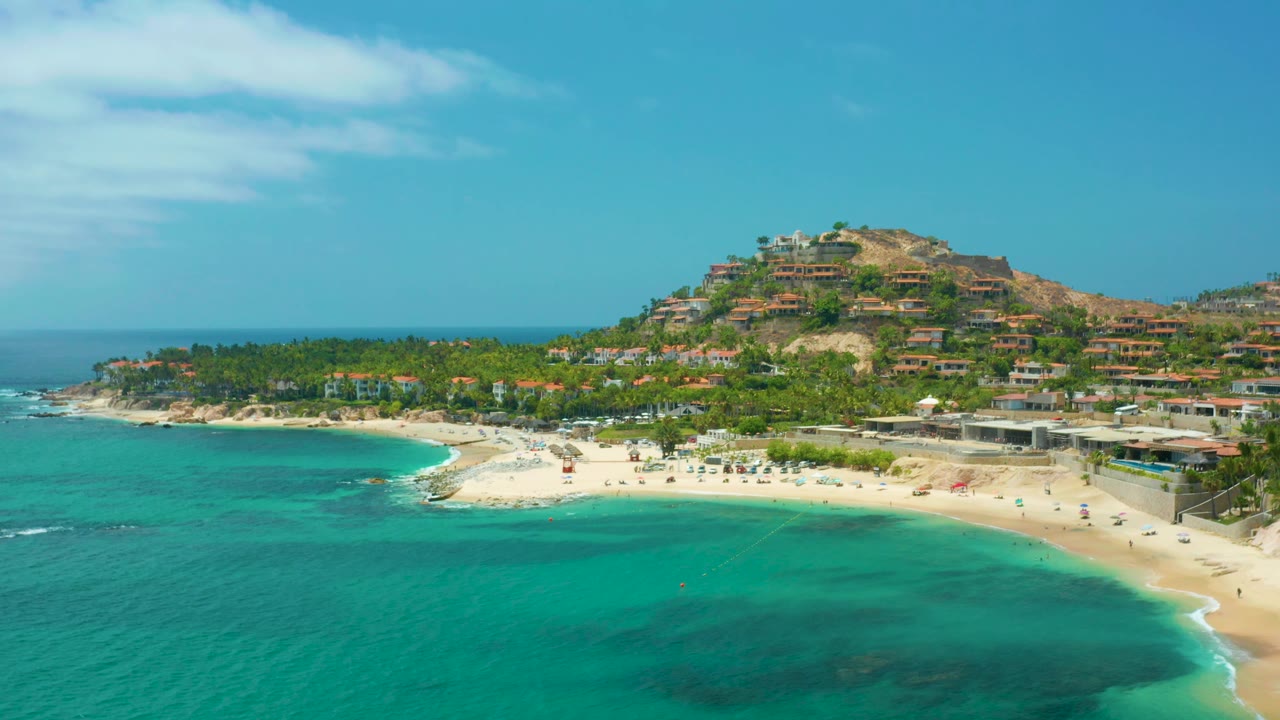 A wide aerial frame of Palmilla Beach Cabo, Mexico showing the bay from slightly farther right, with the beach arc, palm grove, hillside homes, and no visible people.
