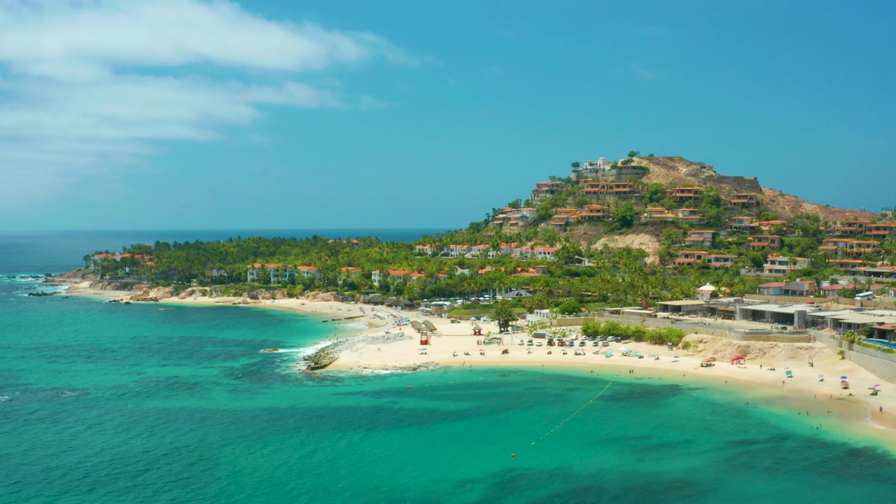 Opening aerial view over Palmilla Beach Cabo, Mexico, with turquoise water in front, a curved sandy beach, hillside homes beyond the palms, and no visible people.