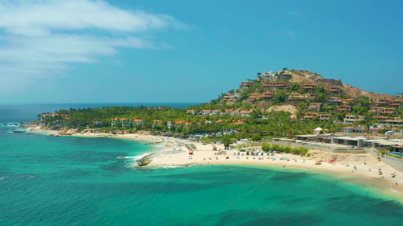Wide drone view of beach, clear water, palms, and hillside housing