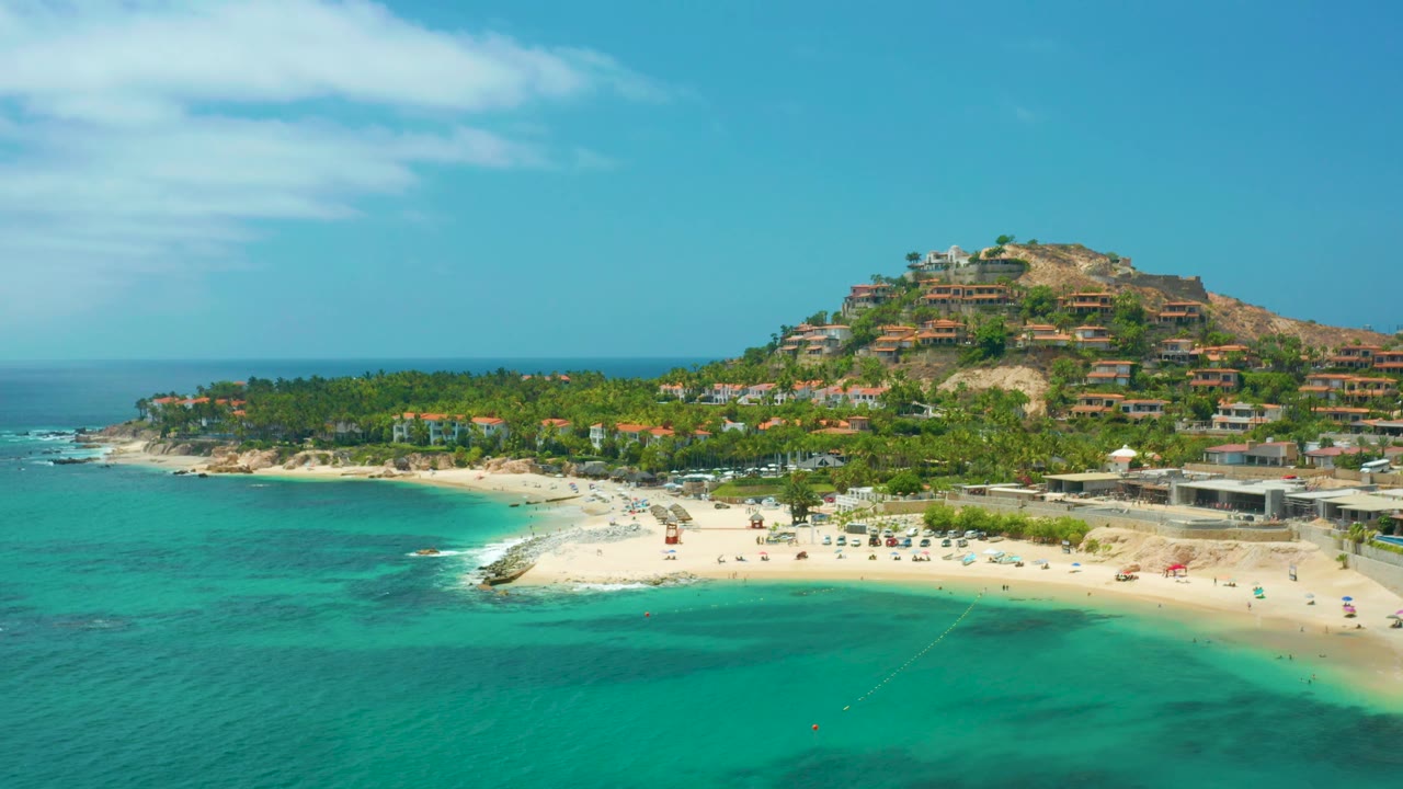 Aerial coastal view at Palmilla Beach Cabo, Mexico, with turquoise sea across the lower frame, beach umbrellas or small beach fixtures along the sand, hillside homes, and no visible people.