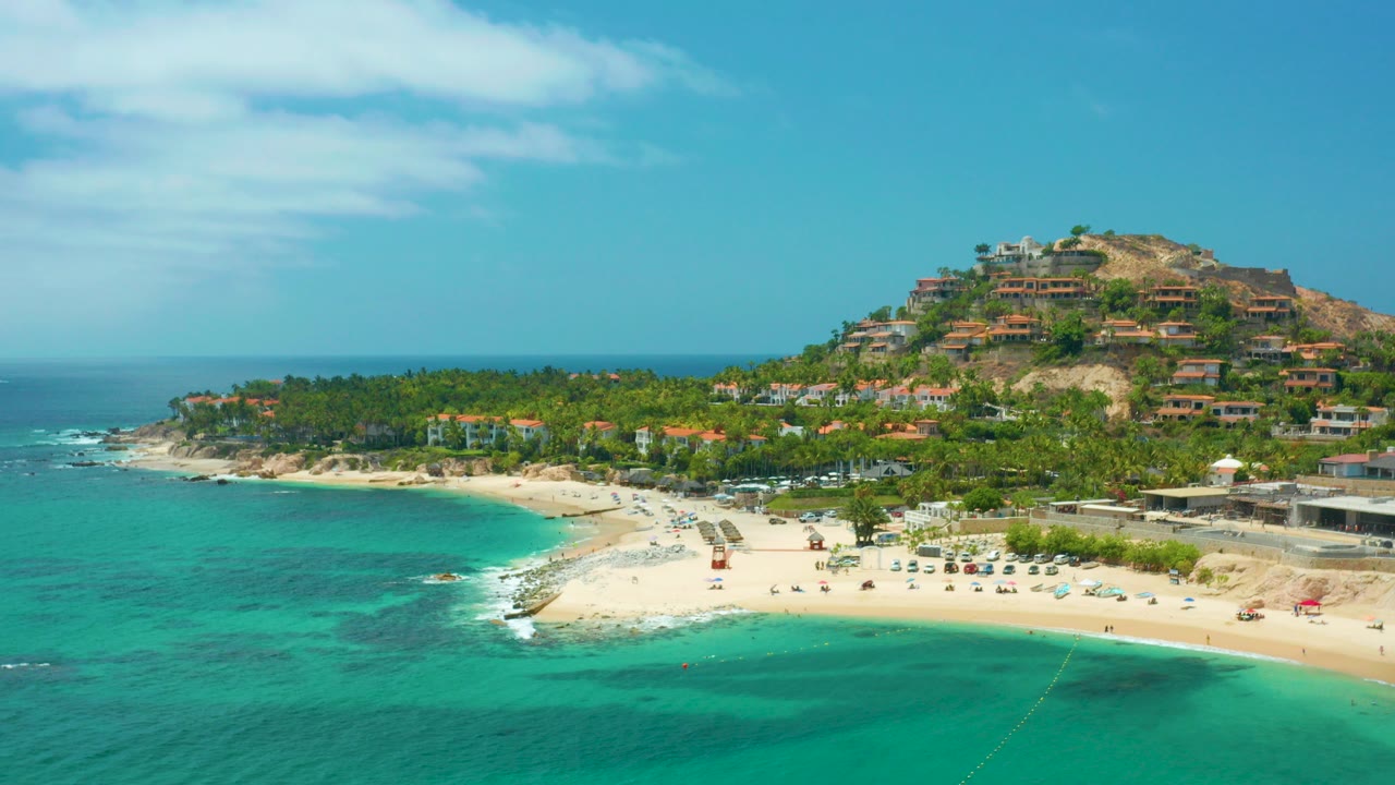 Opening aerial frame over Palmilla Beach Cabo, Mexico, with turquoise water in the foreground, a pale curved beach, palms, hillside buildings, and no visible people.