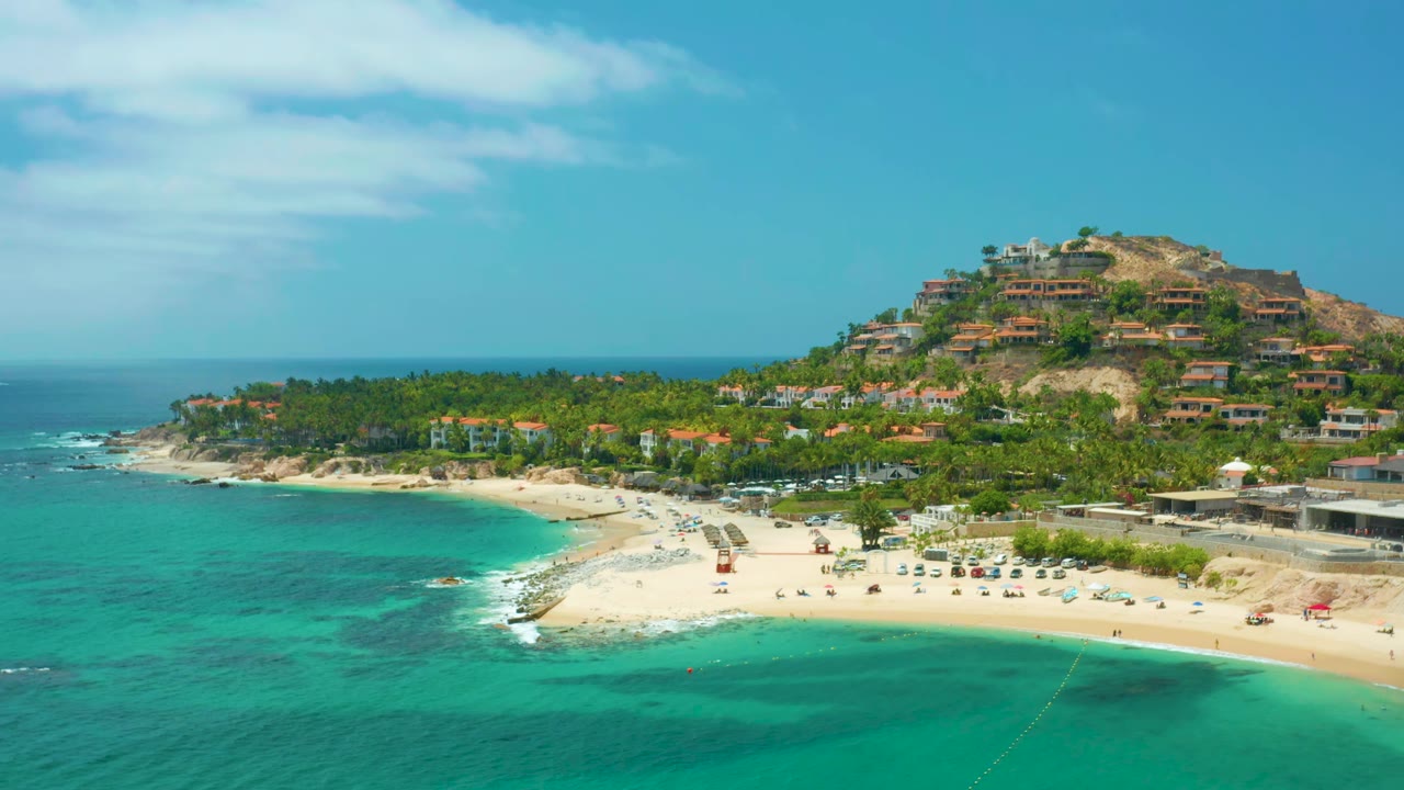 Wide aerial frame at Palmilla Beach Cabo, Mexico, showing shallow blue-green water, a sandy beach with small beach setups, palms, hillside buildings, and no visible people.