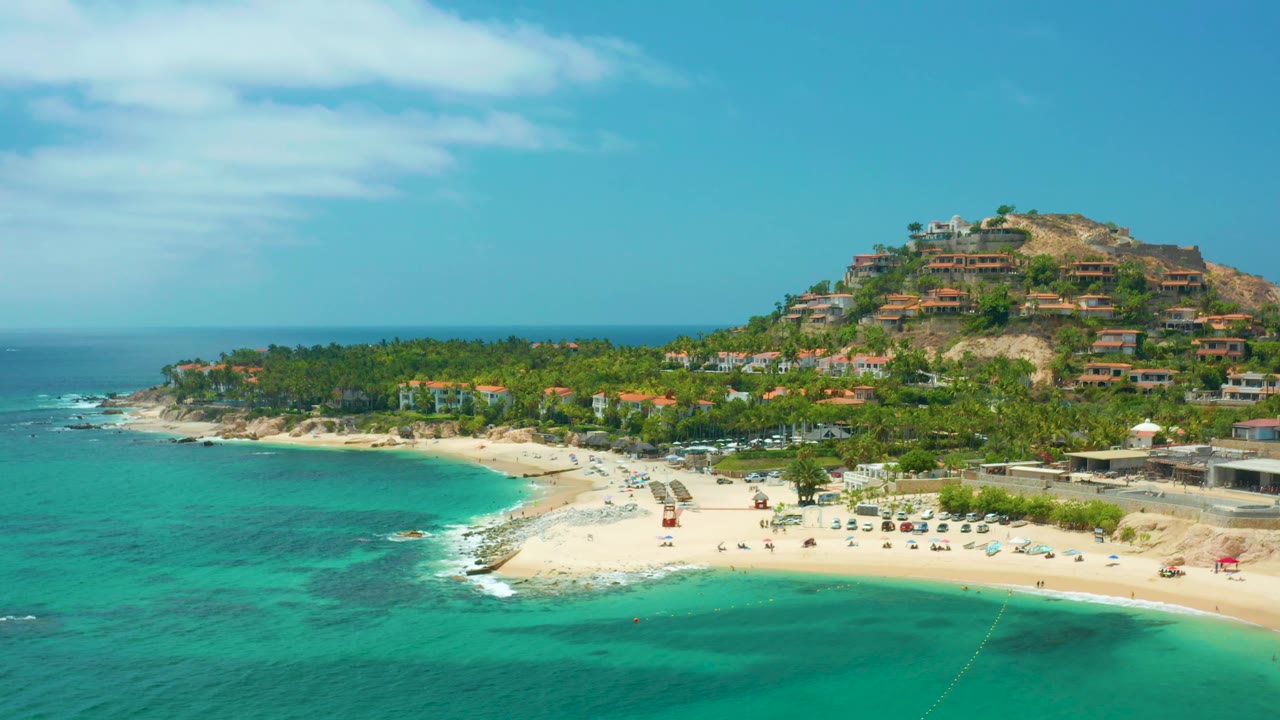 Palmilla Beach Cabo, Mexico aerial frame with open sky above a palm-lined shore, hillside buildings to the right, turquoise water below, and no visible people.