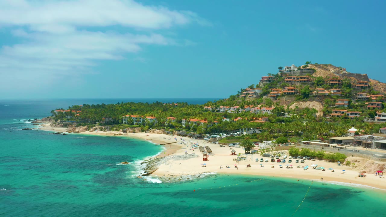 Aerial beach view with turquoise bay, sandy point, palms, and hillside structures.