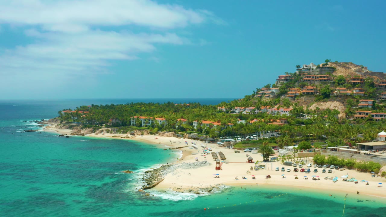 Opening aerial frame of Palmilla Beach Cabo with turquoise water in the foreground, a sandy beach with small visible people, palm trees, and hillside homes under blue sky.