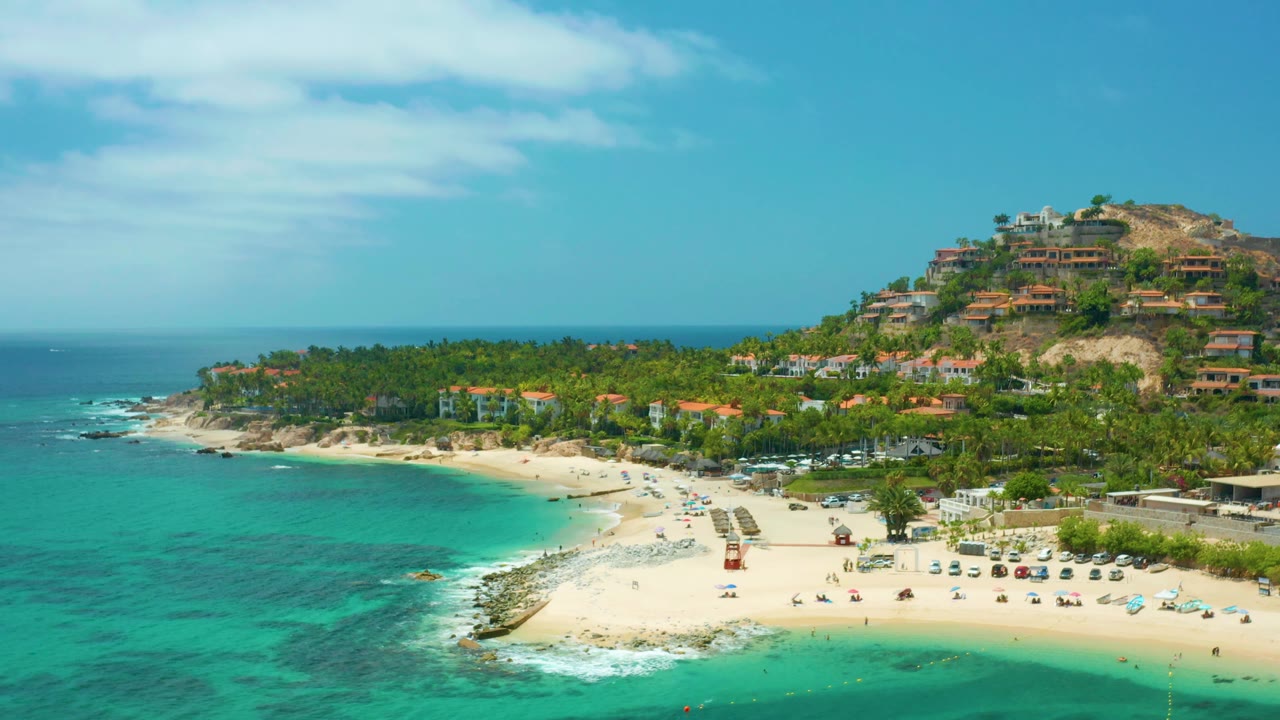 Palmilla Beach Cabo appears from above with the beach centered, clear blue-green water at left, visible people and loungers on the sand, and hillside homes rising at right.
