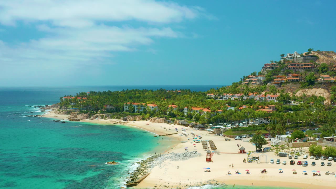 Opening aerial frame of Palmilla Beach Cabo, Mexico showing turquoise water at left, a pale sand beach below, palms across the shore, hillside buildings at right, and no visible people.