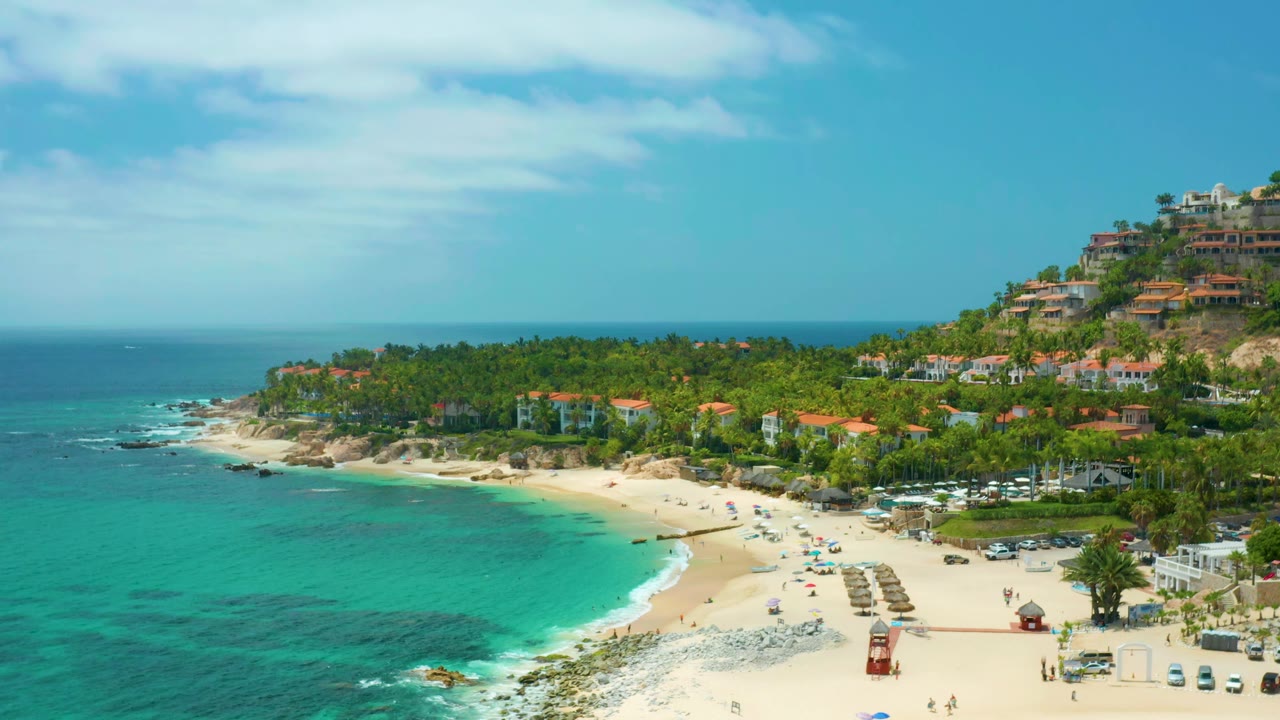 Wide drone view of a curved beach with clear water, palms, and coastal buildings.
