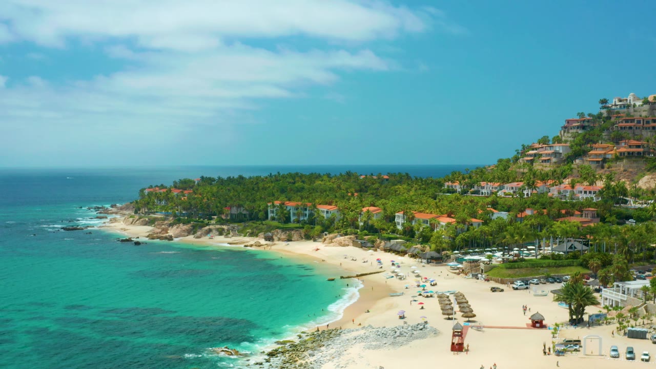 Aerial frame at Palmilla Beach Cabo showing blue-green water in the foreground, a busy-looking beach layout of umbrellas and shade structures, dense palms behind it, and no recognizable people.