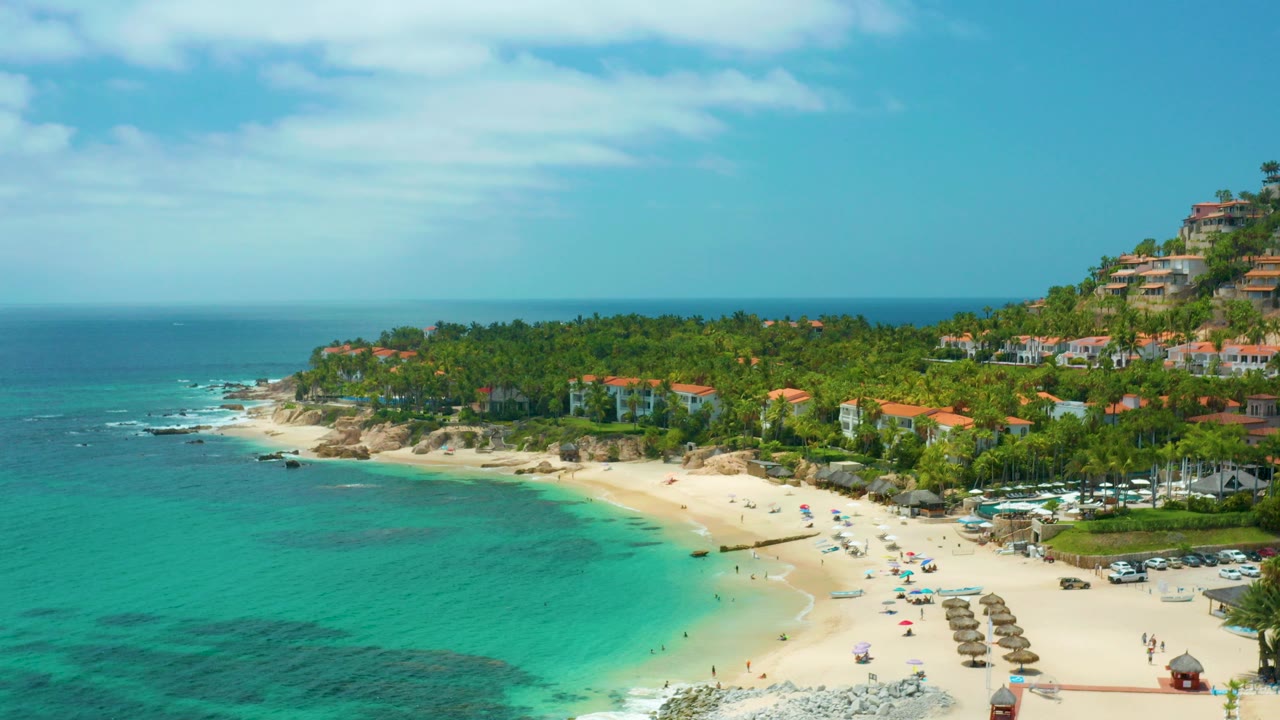 Opening frame of Palmilla Beach Cabo, Mexico from above, with turquoise water in the foreground, pale sand and umbrellas below, hillside resort buildings at right, and no clearly visible people.