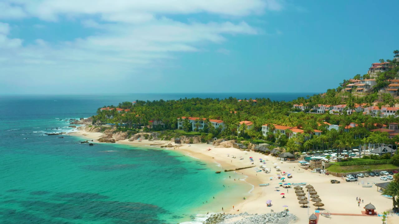 Second frame over Palmilla Beach Cabo, Mexico shows the curved beach and umbrella rows more centrally, with shallow turquoise water at left, palm trees behind the sand, and no clearly visible people.