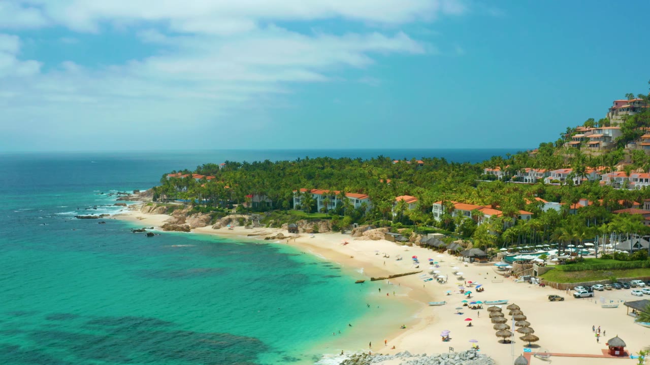 At 0:02, the aerial view of Palmilla Beach Cabo, Mexico keeps the turquoise bay broad in frame, with the sandy resort beach below, palms across the midground, hillside buildings at right, and no clearly visible people.