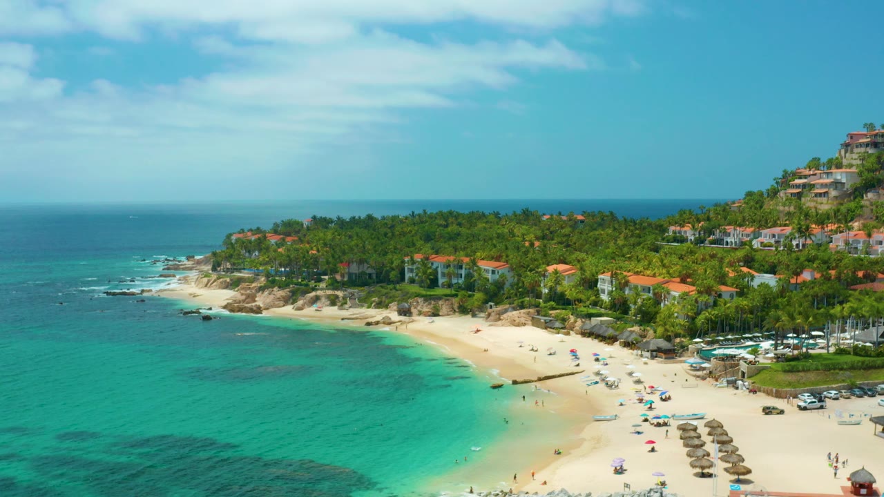 Aerial view of Palmilla Beach with turquoise shallows, sandy beach, palm trees, and hillside lodging.