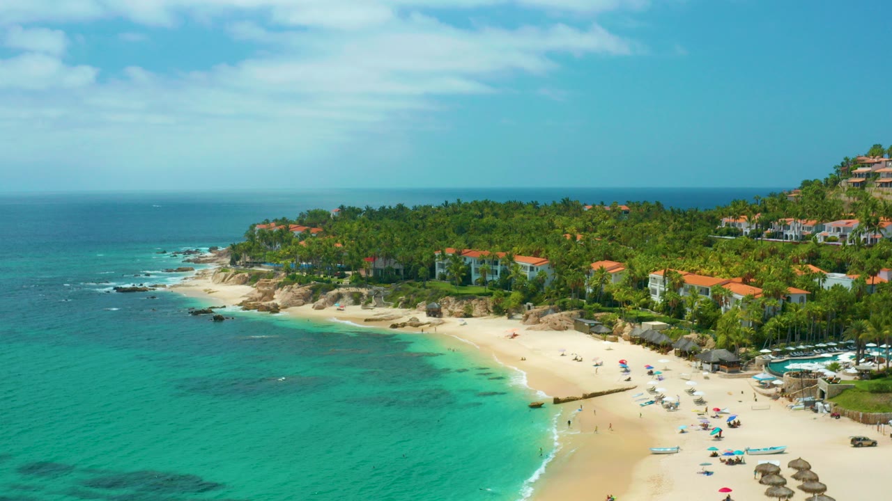 Palmilla Beach Cabo, Mexico from above, with turquoise water in the foreground, a curved sandy beach, small umbrellas, distant beachgoers, and hillside homes under a blue sky.