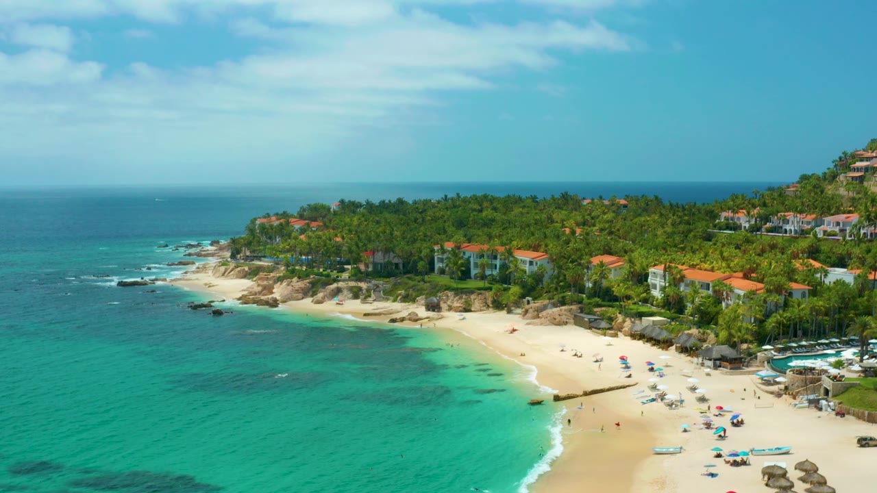 Palmilla Beach Cabo appears from a high angle with open turquoise water at left, a sandy beach at lower right, small visible people, and palm-covered homes along the coast.