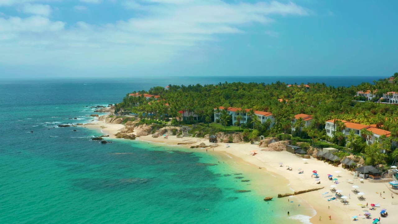 High coastal view with blue-green water, sand, rocks, palms, and homes.
