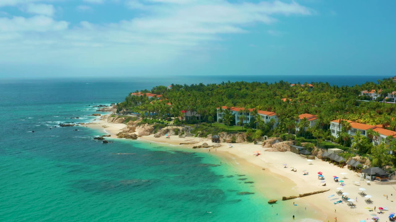 Wide aerial frame of Palmilla Beach Cabo, Mexico, with clear water along the left foreground, pale beach on the right, palms and coastal homes behind, and no visible people.