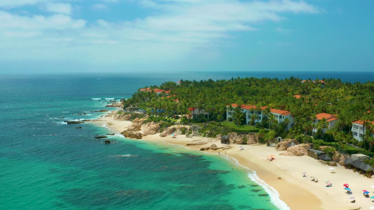 Opening aerial frame over Palmilla Beach Cabo, Mexico, with turquoise sea in the foreground, a curved sandy beach, rocky shoreline, palm trees, red-roofed buildings, and no visible people.