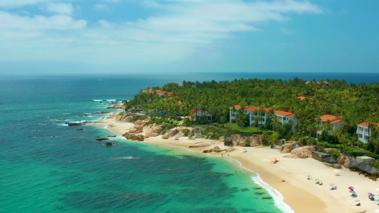 Wide aerial coastline with turquoise surf, pale sand, palms, rocks, and red-roofed buildings.