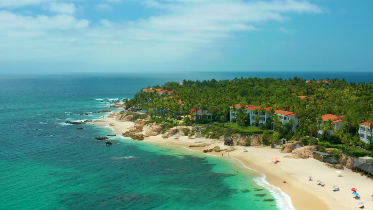 At 0:02, Palmilla Beach Cabo fills the frame with clear turquoise shallows, a bright sand strip, scattered rocks, palm greenery, red-roofed coastal buildings, and no visible people.