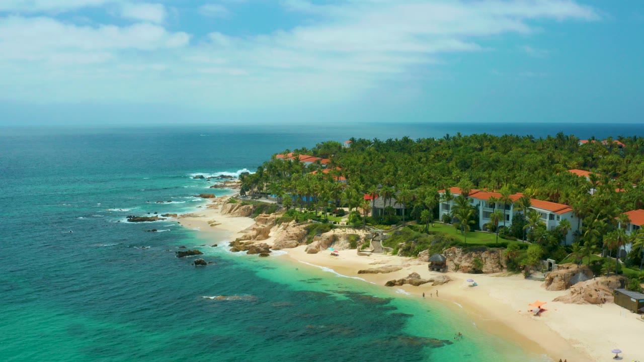 Aerial view over Palmilla Beach Cabo, Mexico with turquoise water at left, pale sand below, rocky shore, red-roofed villas among palms, and no visible people.