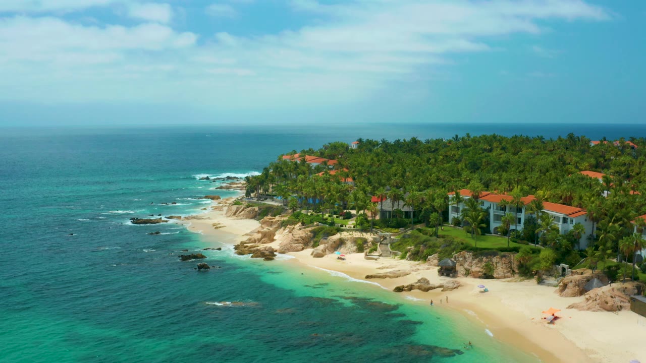 Elevated view of villas and palm trees above the sandy edge of Palmilla Beach Cabo, Mexico, with blue-green water and scattered rocks offshore, and no visible people.