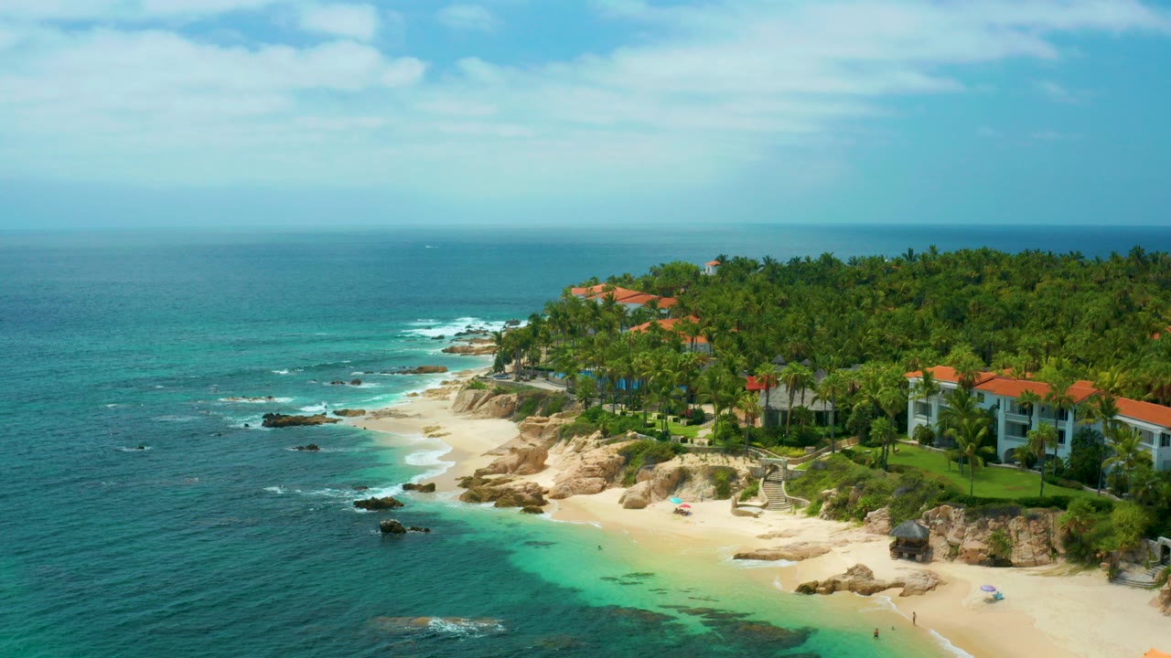 Palmilla Beach Cabo, Mexico aerial frame with turquoise water in front, a sandy cove and rocks along shore, red-roofed homes among palms, and no visible people.
