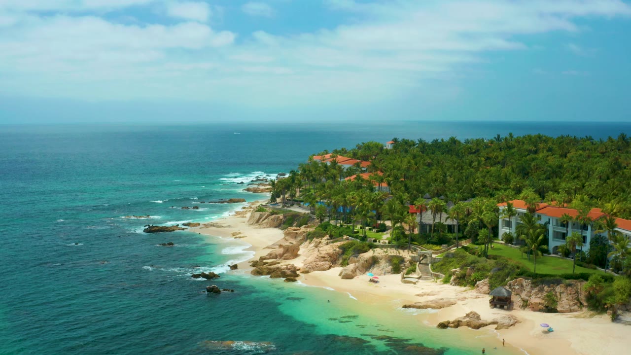 Palmilla Beach Cabo, Mexico frame showing the ocean on the left, waves wrapping around rocks, a bright sand beach, homes behind palms, and no visible people.