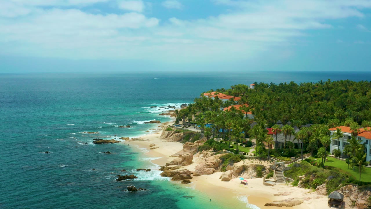 Opening aerial frame over Palmilla Beach Cabo, Mexico, with blue ocean at left, pale sand below, rocky shoreline, palm trees, red-roofed villas, and no visible people.