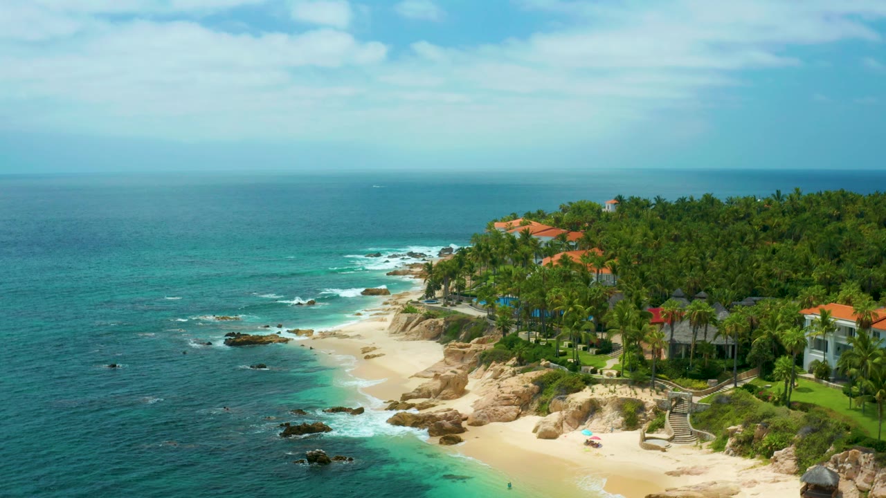 Aerial frame at 0:02 over Palmilla Beach Cabo, Mexico, with the horizon high in frame, open sea to the left, sand and rocks below, villas on the right, and no visible people.