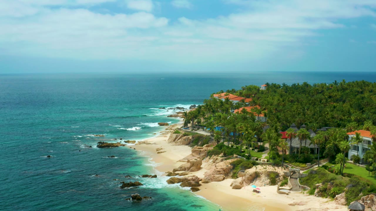 Ocean shoreline with rocks, sand, palms, and villa buildings.