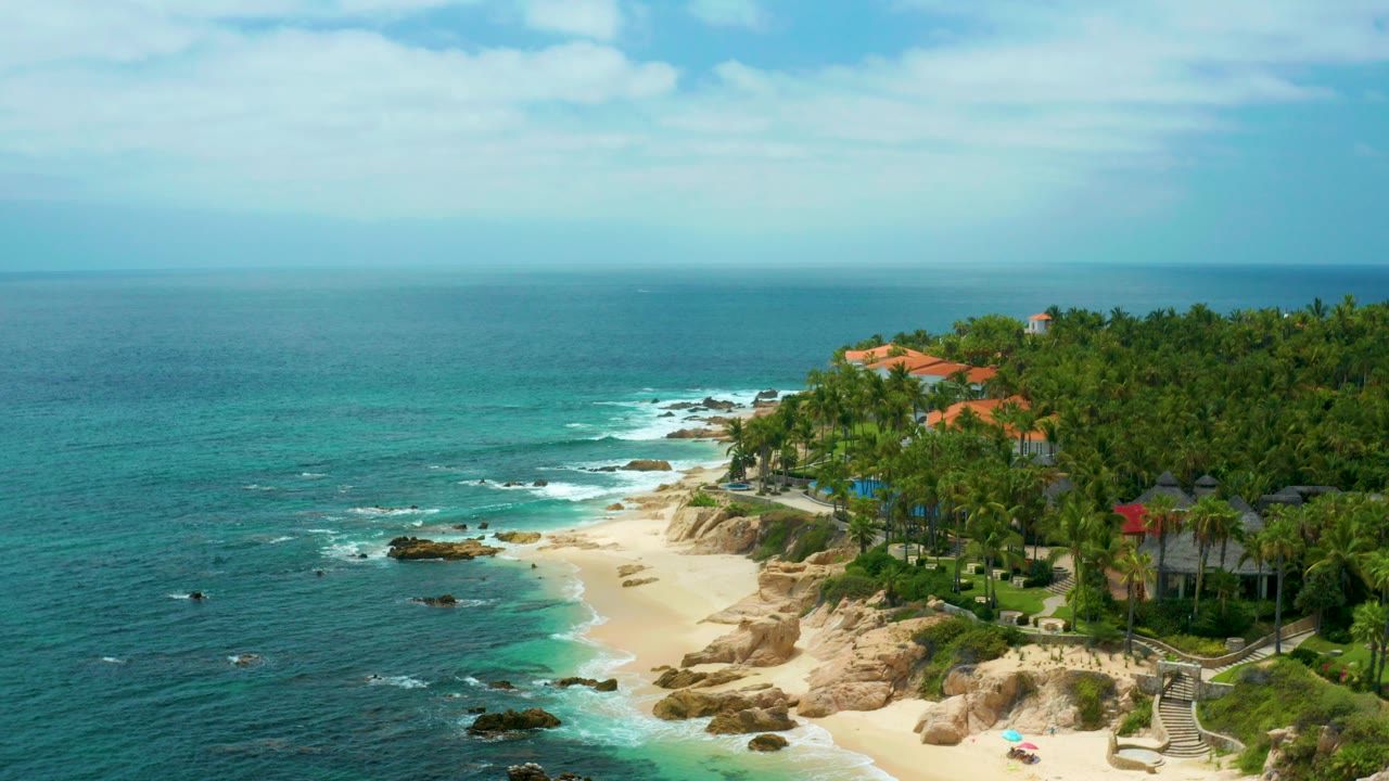 Opening aerial frame over Palmilla Beach Cabo, Mexico, with turquoise water on the left, a sandy cove below, tan rocks, dense palms, red-roofed buildings, and no visible people.