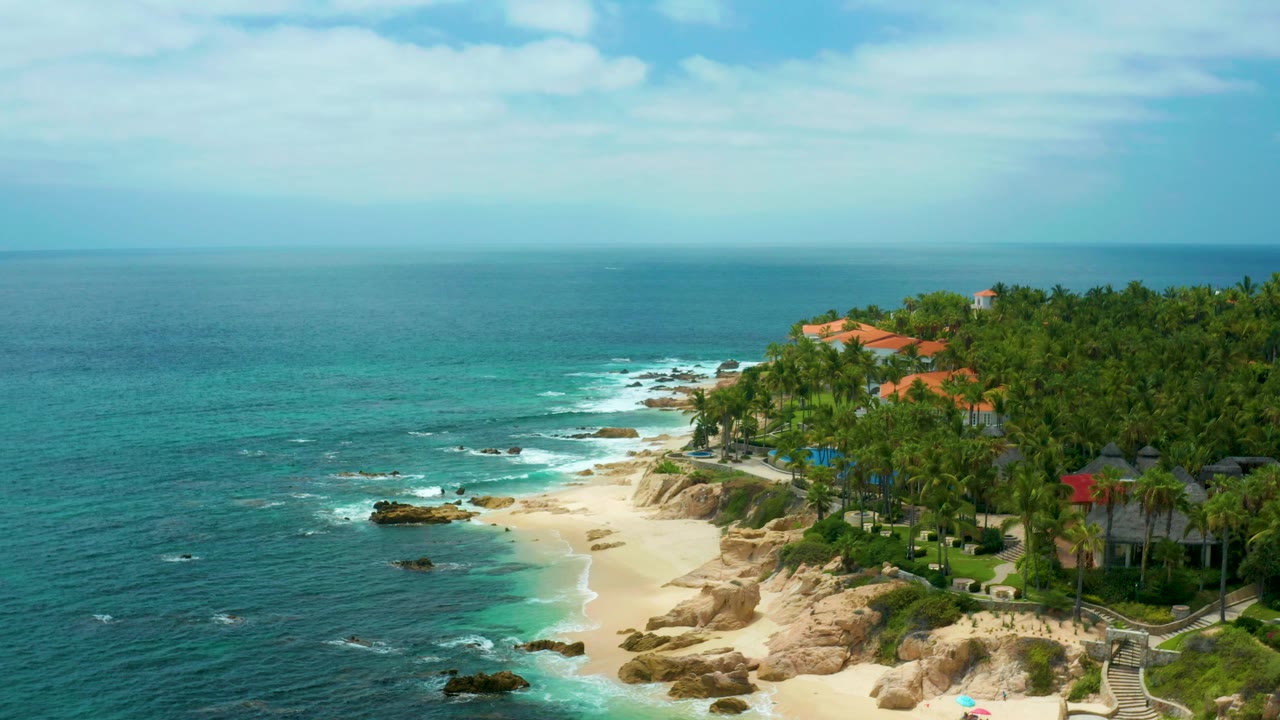 Aerial beach view with open water, small surf, rocks, palms, and hillside buildings.