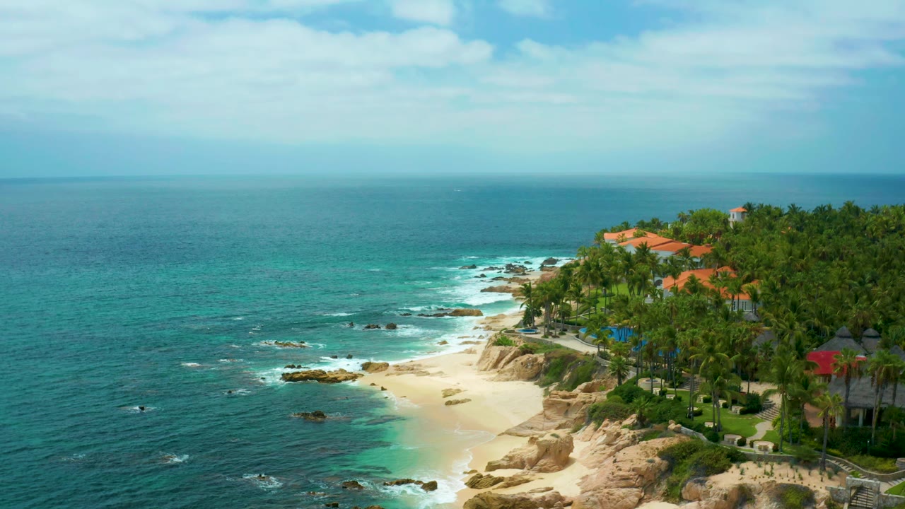 Wide aerial coastline view with surf lines, beach, cliffs, palms, and red roofs.