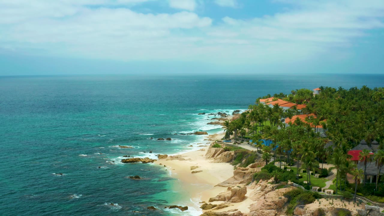 Aerial view over Palmilla Beach Cabo, Mexico with more open water in frame, small waves near rocks, a light sand beach, palm-covered shoreline, rooflines, and no visible people.