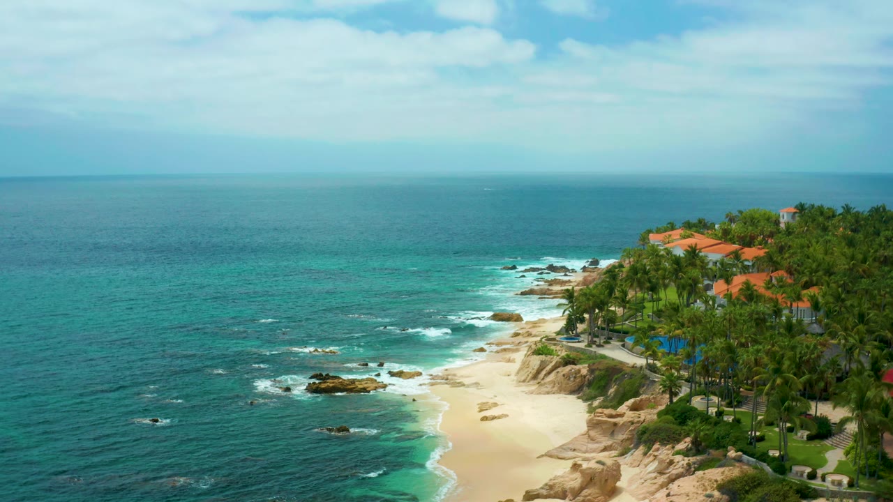 Palmilla Beach Cabo aerial view with blue-green water spread across the left side, small waves on rocks, a bright sand strip, dense palms, orange rooftops, and no visible people.