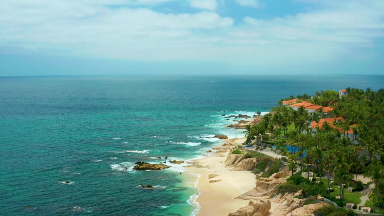 Wide Palmilla Beach Cabo frame showing open ocean across most of the view, foamy water near dark rocks, a sandy beach curve, palm-covered grounds, orange roofs, and no visible people.