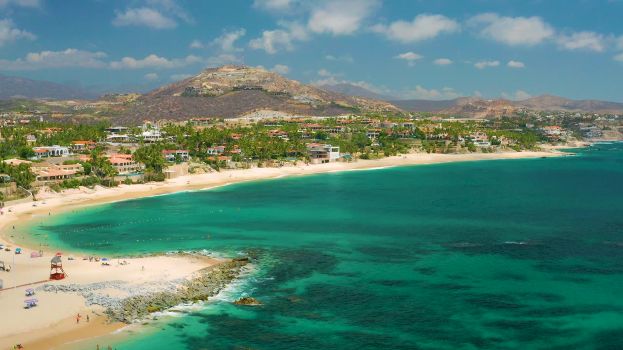Palmilla Beach Cabo, Mexico appears from above with a rocky foreground point, pale sand, turquoise water, waterfront buildings, dry hills, and tiny people visible near the lower beach.