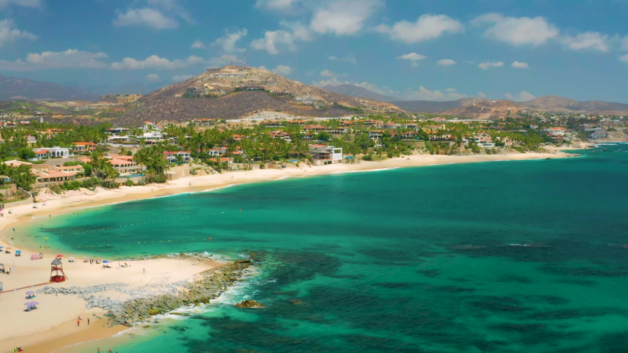 High view over a curved beach with green-blue water and shoreline buildings.